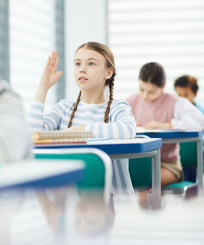 Vertical portrait of young schoolgirl sitting and desk raising hand and asking question to her teacher during lesson time, copy space